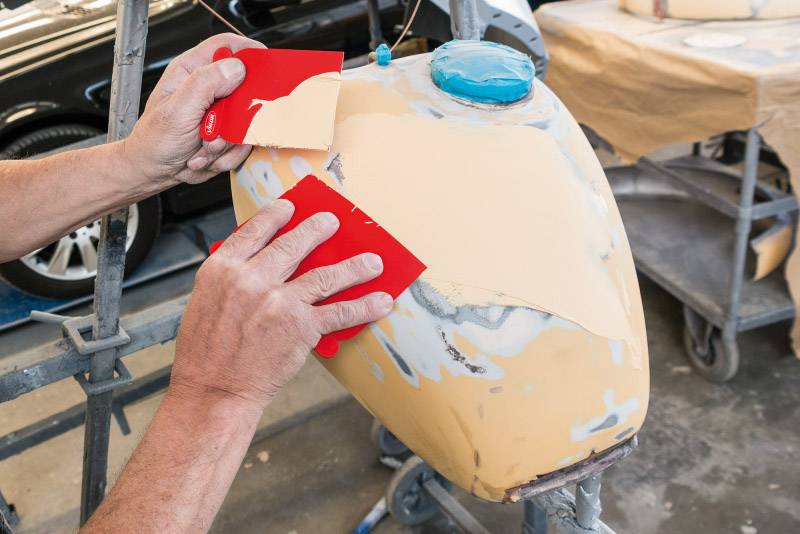 A person is working on a sand-coloured tank with red sanding blocks in a workshop environment.