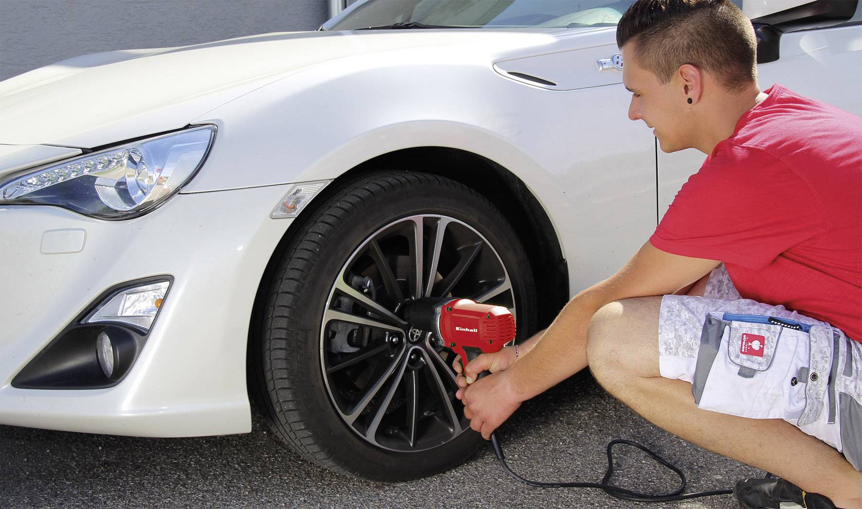 A man is changing the tyre of a white car using an electric tool. A concrete wall is visible in the background.