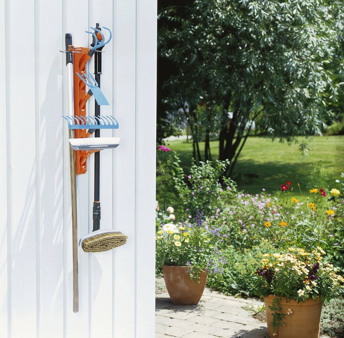 Garden tools mounted on the wall beside a flowering garden with a stone path and potted plants in the foreground.
