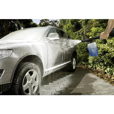 A car is being sprayed with a pressure washer outdoors. Foam covers the surface, while water sparkles in the sunlight.