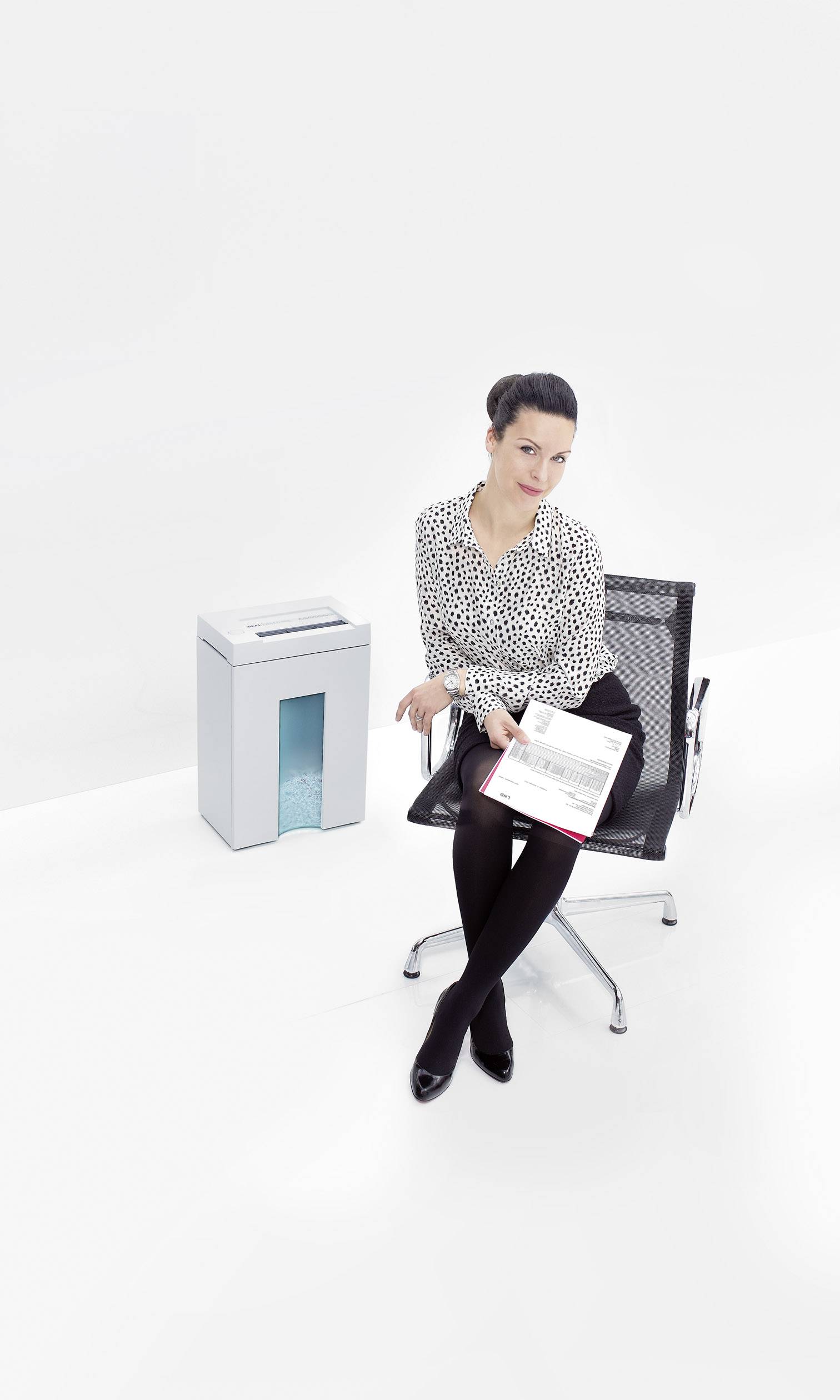 A woman is sitting on an office chair next to a paper shredder, holding documents in her hand. The background is plain white.