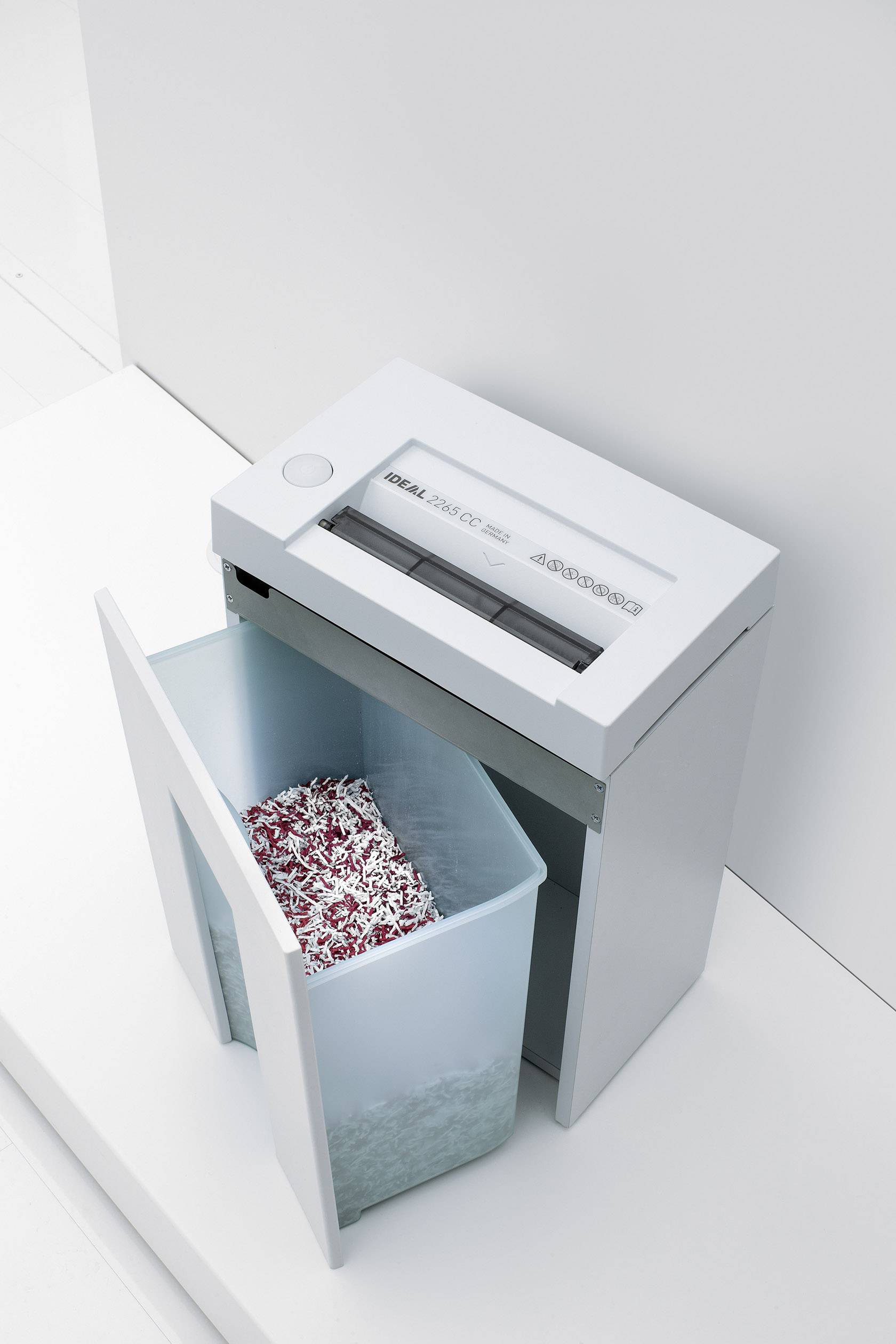 A paper shredder with an open paper collection bin, filled with shredded paper pieces. Device is standing on a white floor.