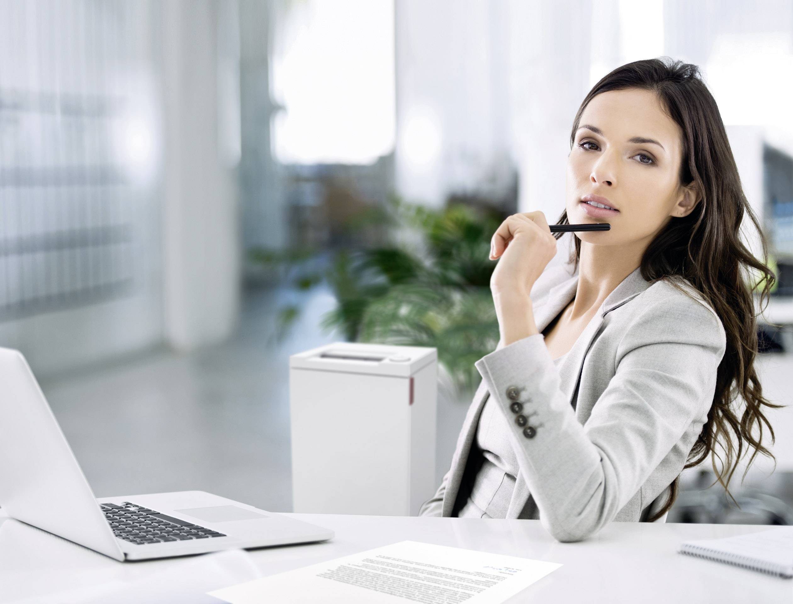 A woman in a grey suit sits thoughtfully in front of a laptop, pen in hand, in a modern office.