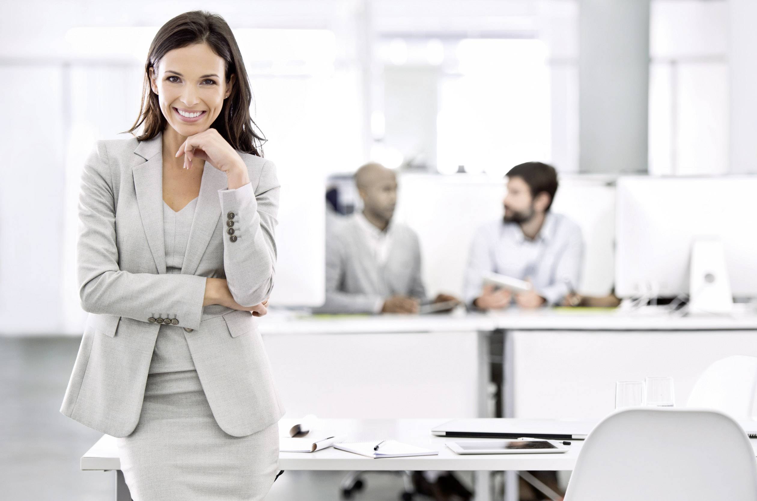 A smiling woman in business attire is standing at the front of a modern office. In the background, two people can be seen at a desk.