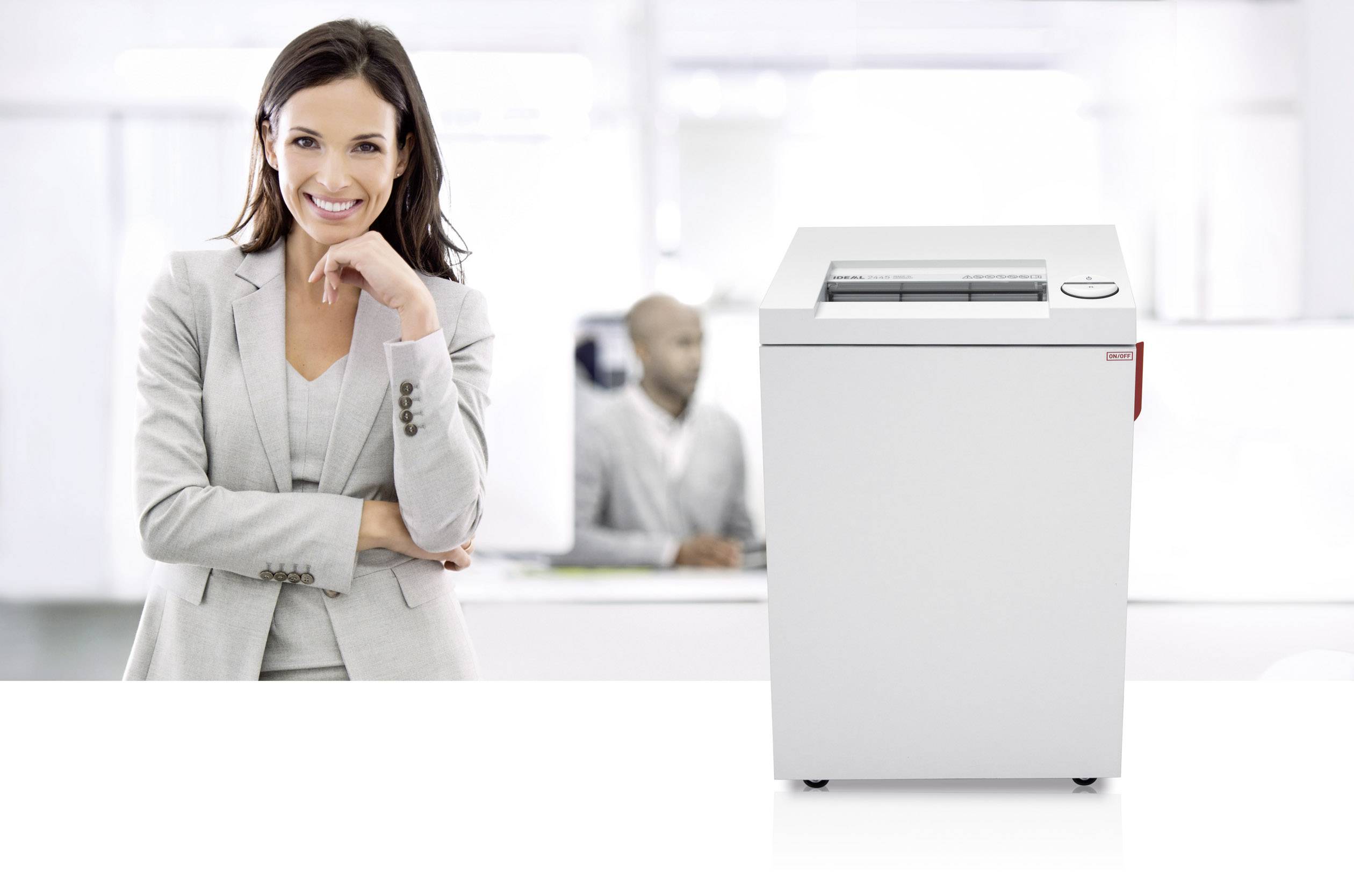 A woman in a grey suit stands smiling next to a paper shredder in an office.