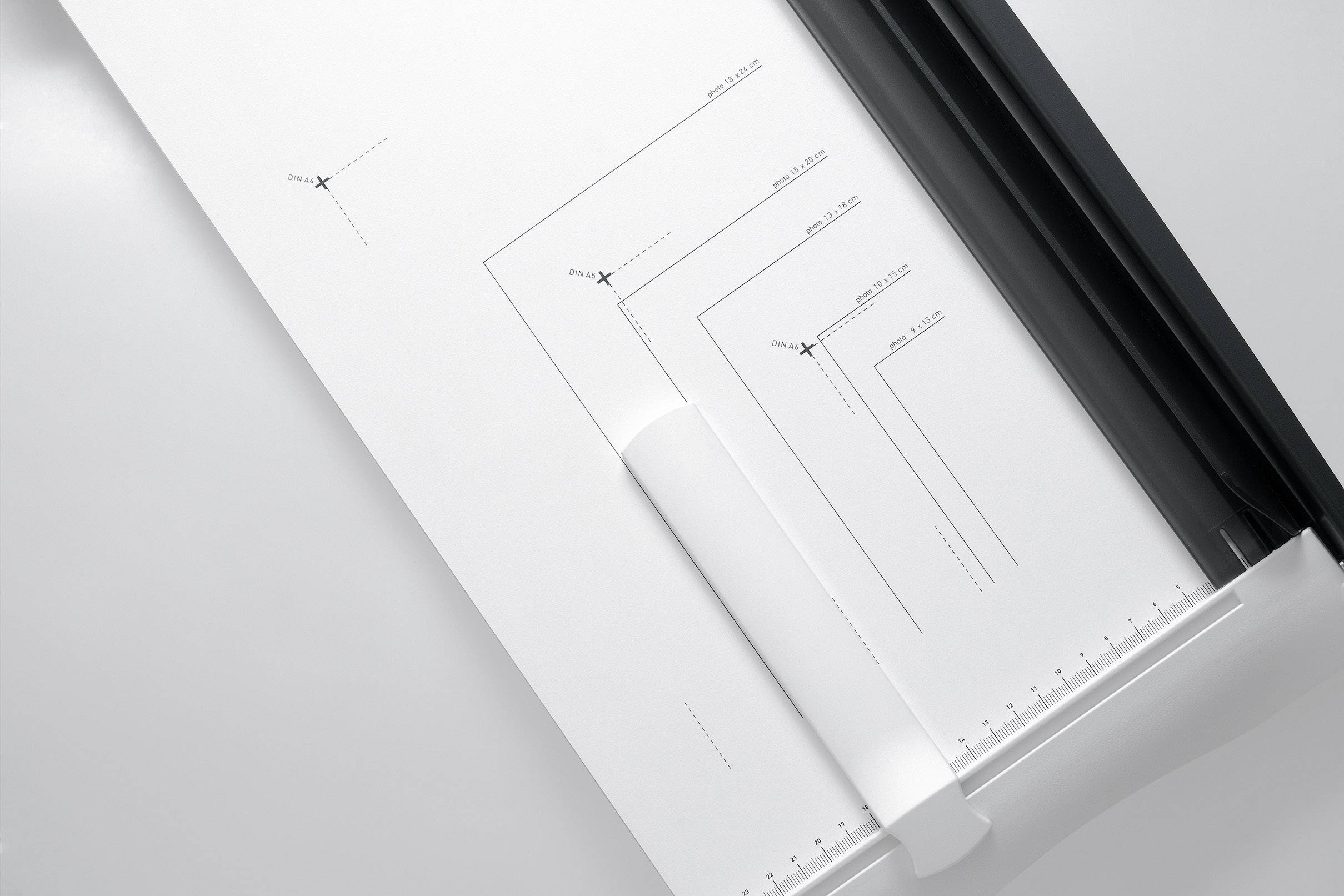 A paper guillotine cuts white paper. The paper has lines and measurement markings. Primary function: precise cutting.