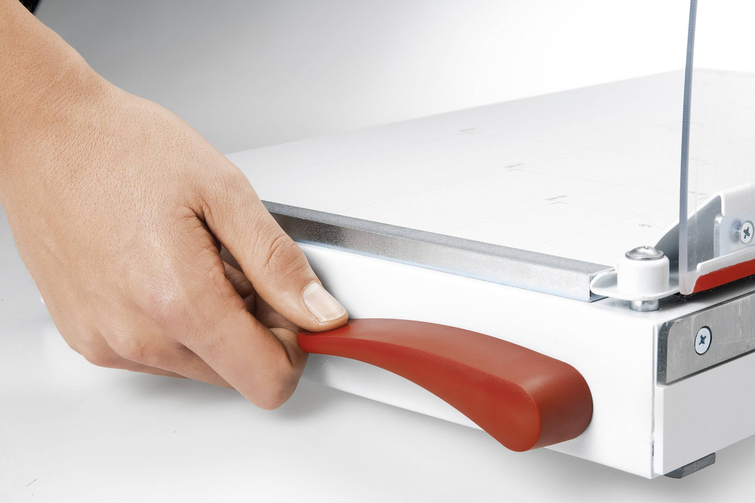 A hand operates the red lever of a paper cutter. The lever is attached to the underside of a metal cutting device.