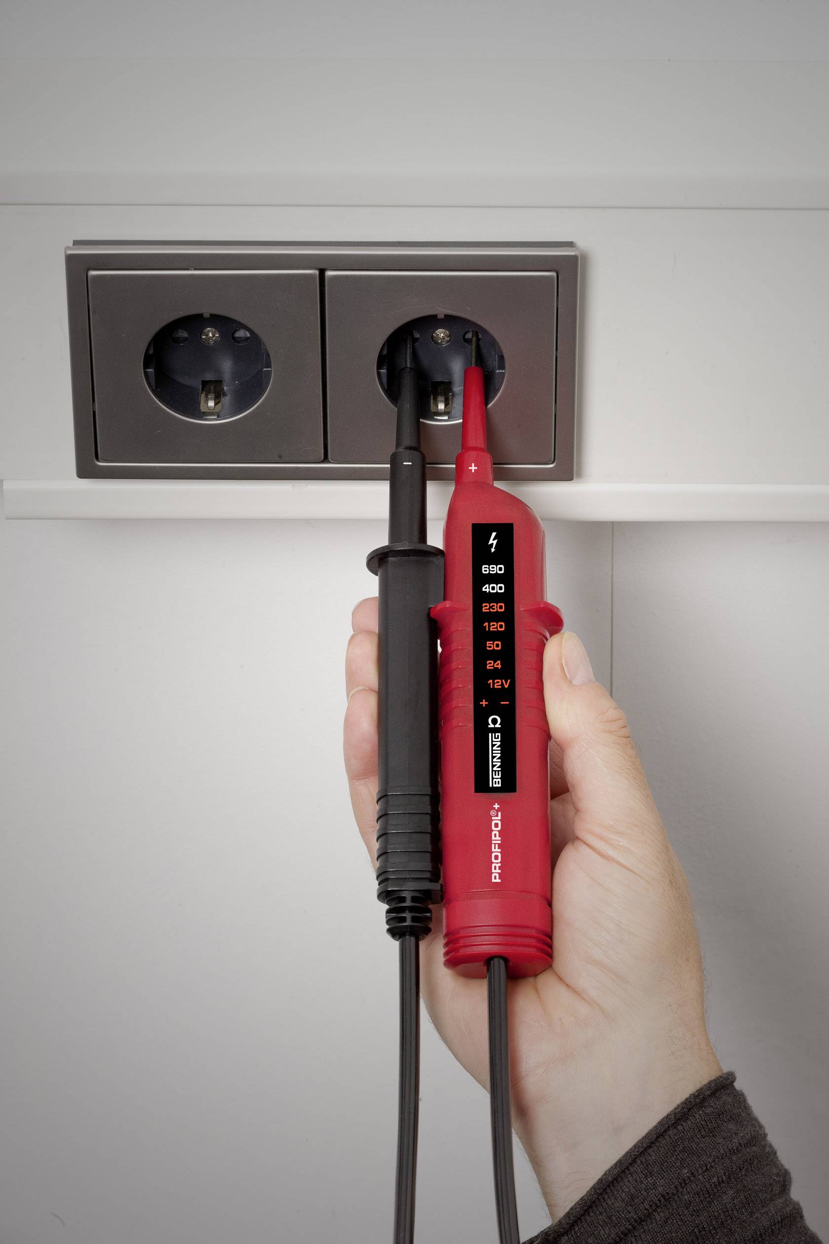 A hand is holding a two-pole voltage tester against a power socket to test the electrical current. The device is displaying measurement readings.