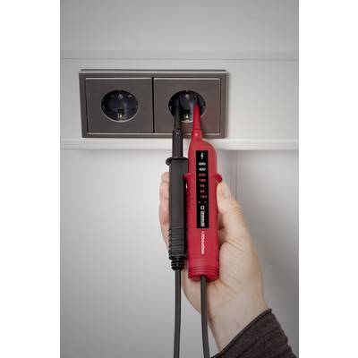 A hand is holding a two-pole voltage tester against a power socket to test the electrical current. The device is displaying measurement readings.
