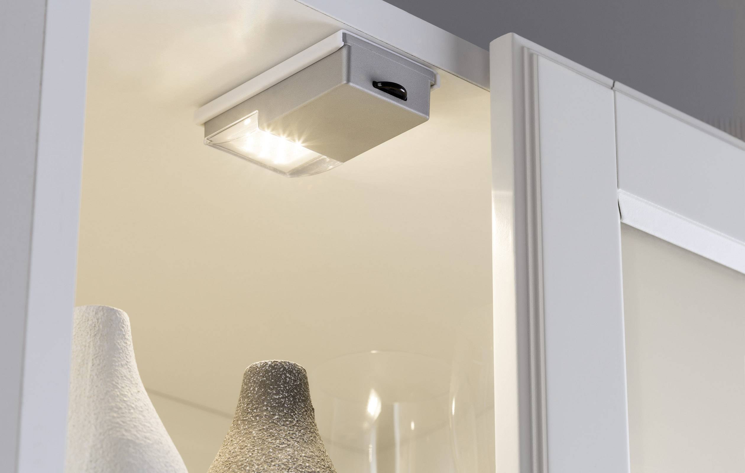 Interior cabinet lighting shows an LED spotlight illuminating white ceramic vases in an open cupboard, providing bright, directional light.