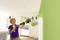 A person is spraying green paint onto an interior wall using a paint spraying system in a bright room.