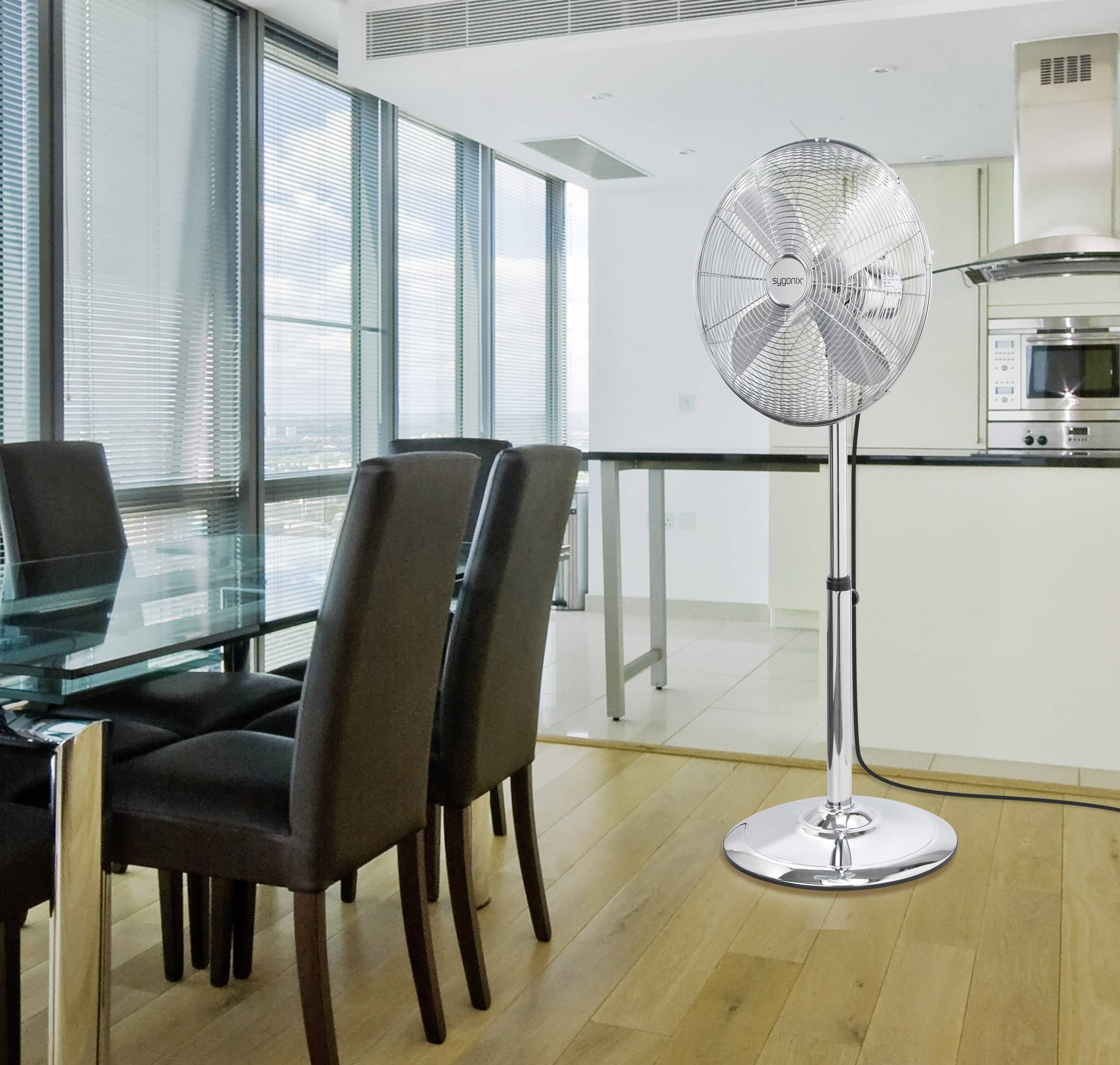 Modern dining area with a glass table and black chairs, adjacent to an open-plan kitchen. A silver pedestal fan in the room.