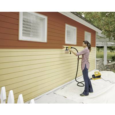 A woman is painting the exterior wall of a house with a paint sprayer. The upper wall is rust red, the lower section beige.
