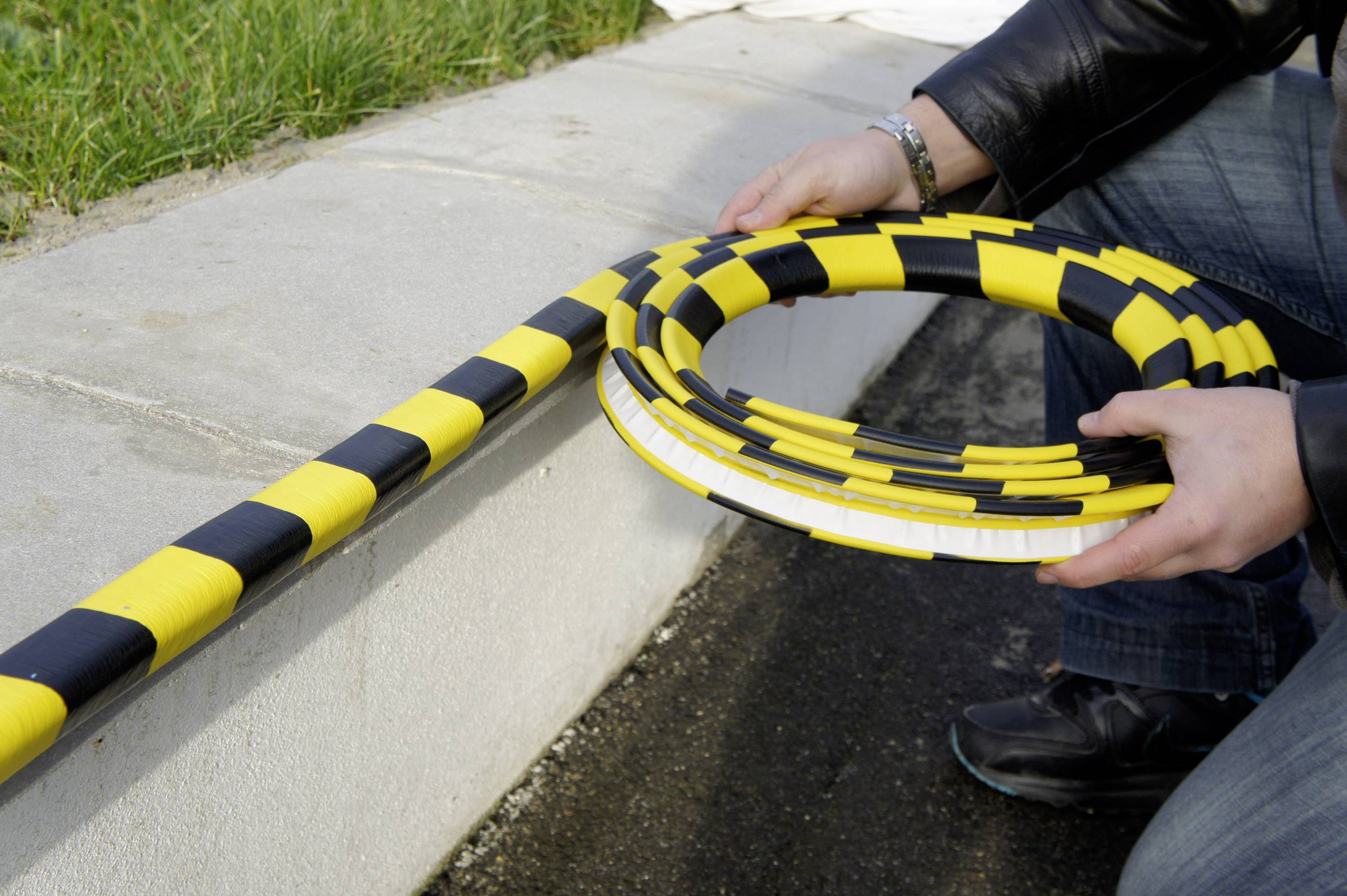 A person is unrolling a yellow and black hazard tape along a kerb edge to mark off an area.