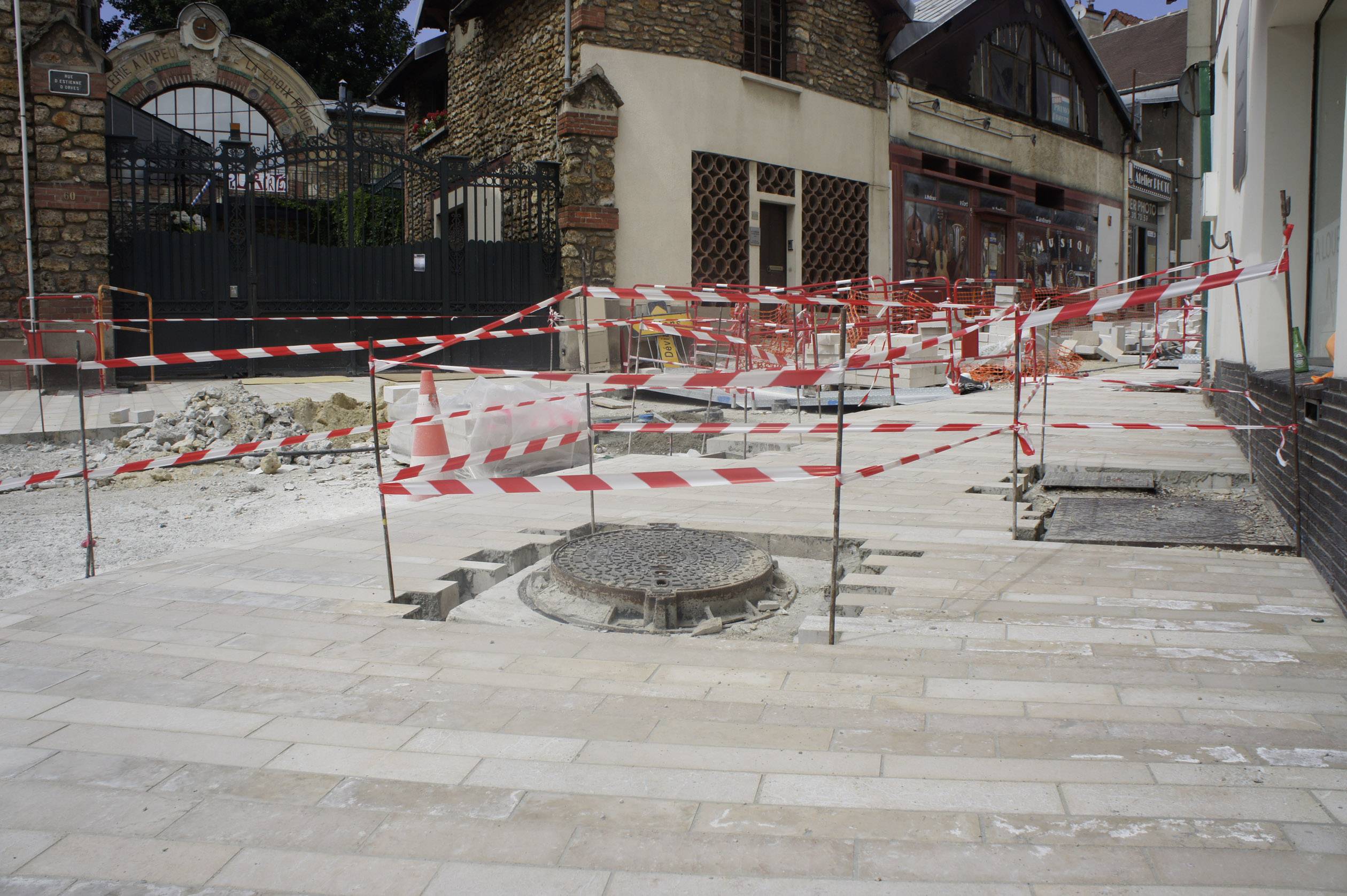 Road works on a paved street, surrounded by red and white barrier tape. No workers are visible. Buildings can be seen in the background.