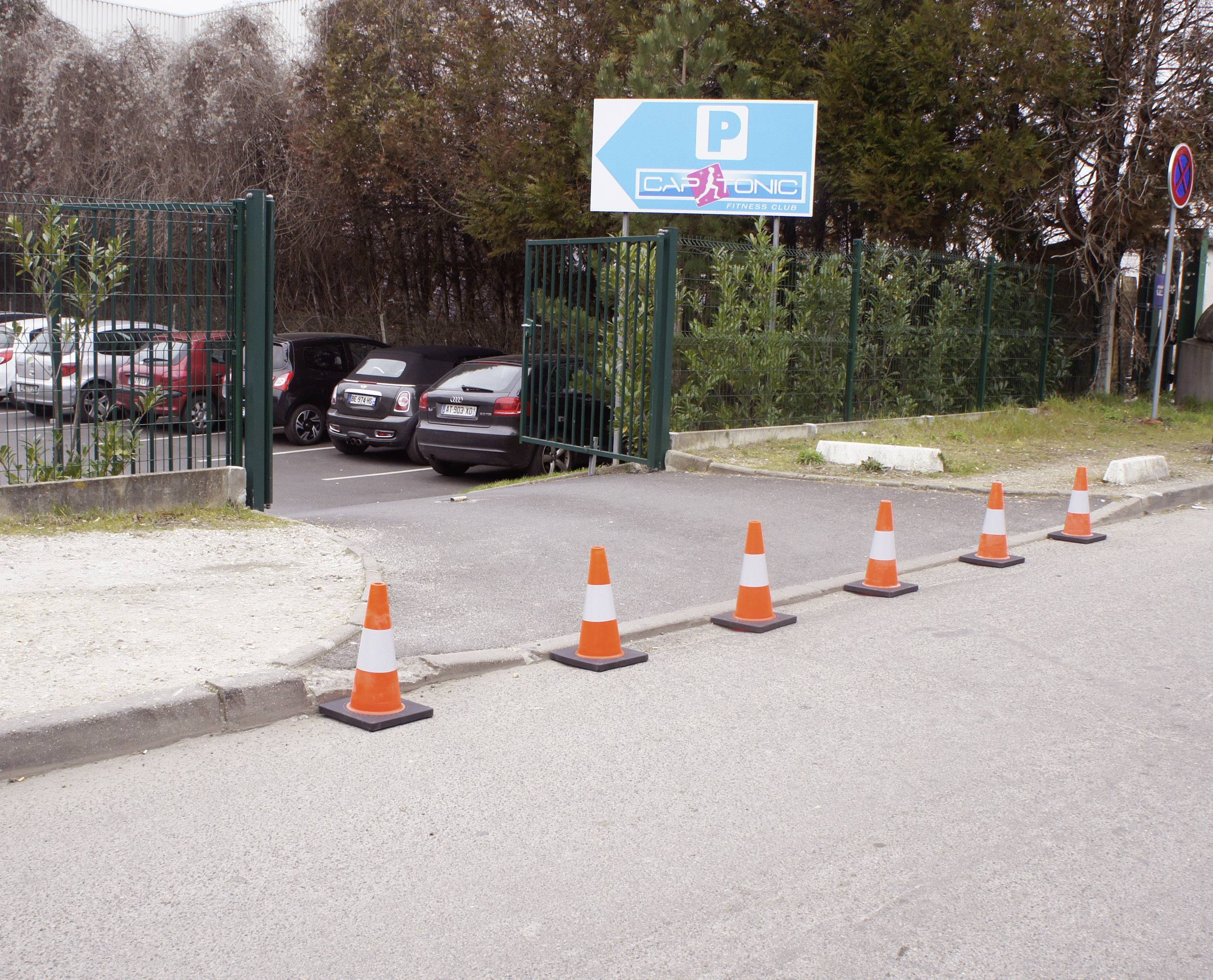 Cars drive through an open gate into a car park. A blue sign with 'Parking' shows the way. Traffic cones are positioned at the edge.