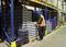 A worker wearing a high-visibility orange safety vest is stacking grey plastic crates in a warehouse with tall shelving units filled with cardboard boxes.