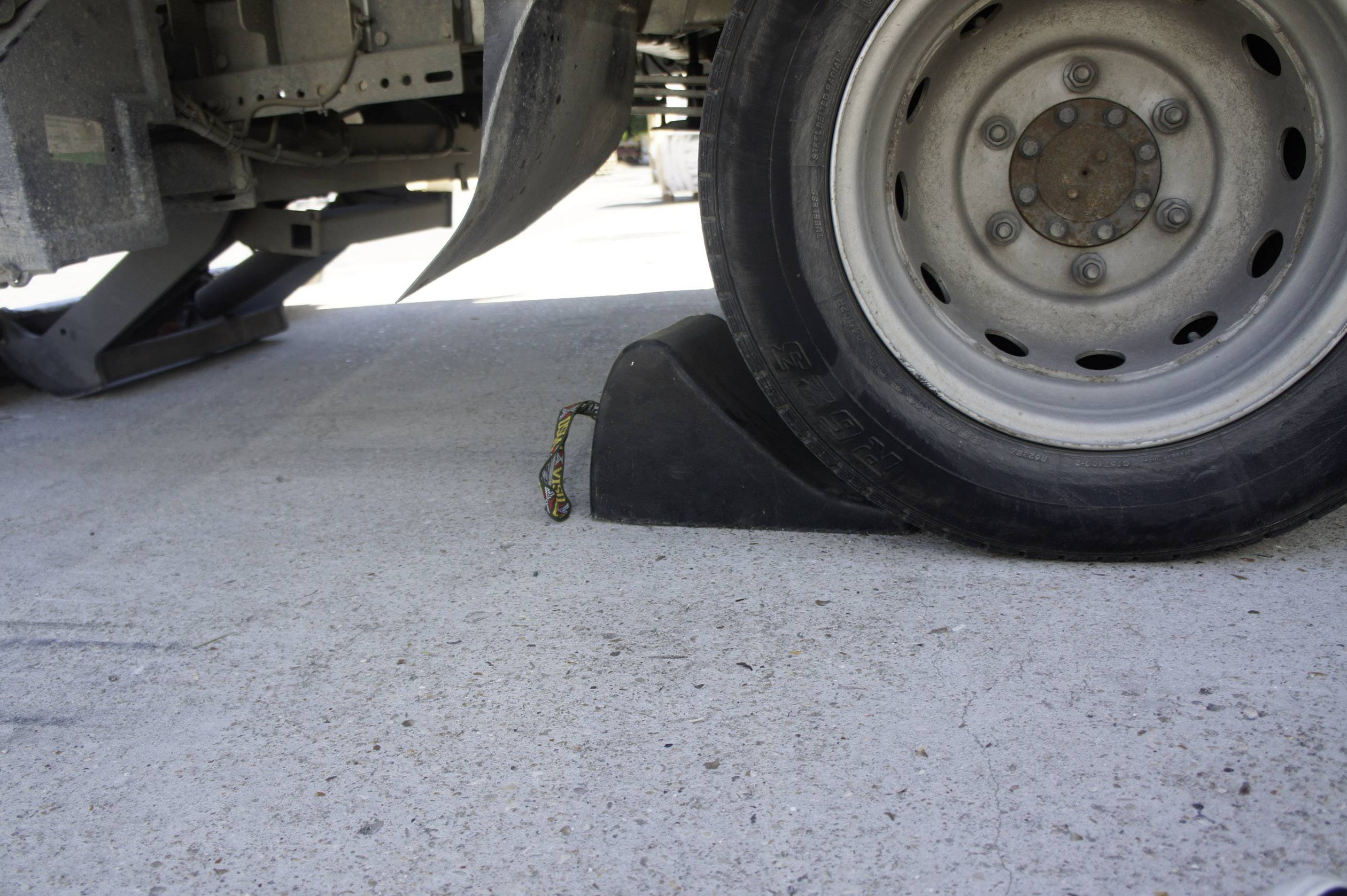 A lorry tyre is secured with a wheel chock. The chock prevents the vehicle from rolling away on a flat concrete surface.