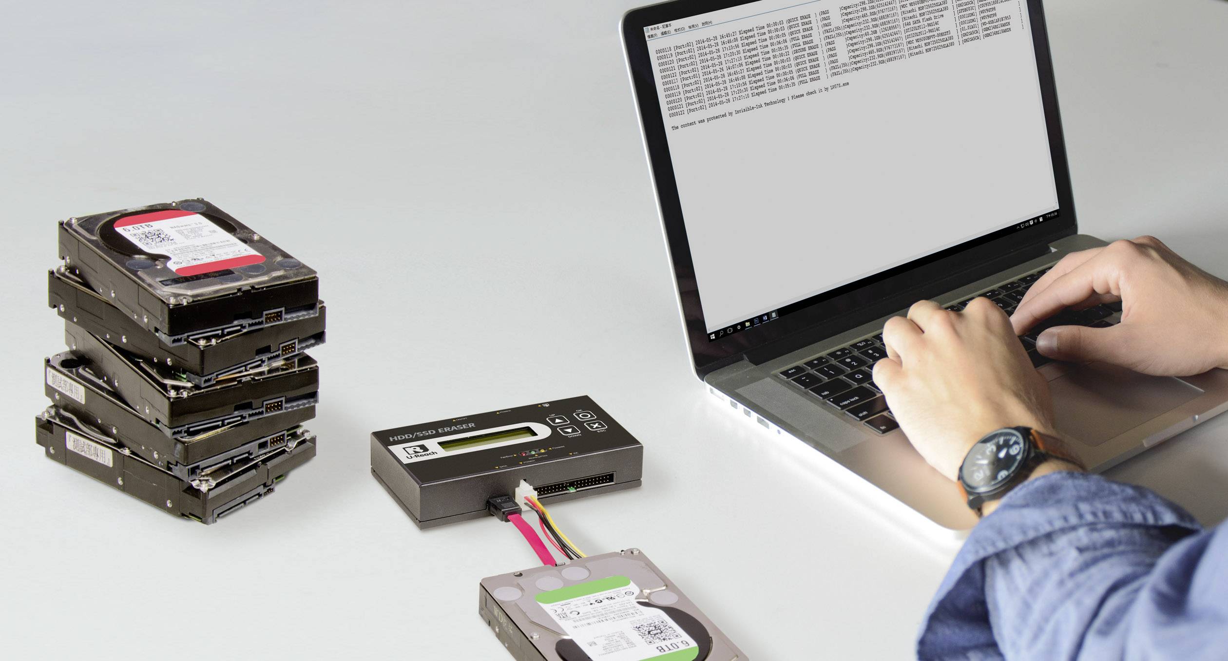 A person is working on a laptop next to a stack of hard drives and a data erasure device. Data erasure is displayed on a screen.