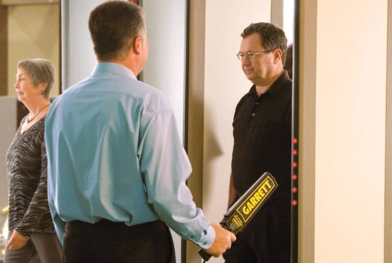 A man is being checked at a security checkpoint with a handheld scanner; another person walks past.