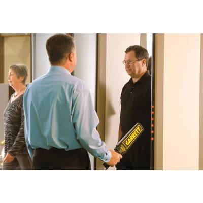 A man is being checked at a security checkpoint with a handheld scanner; another person walks past.