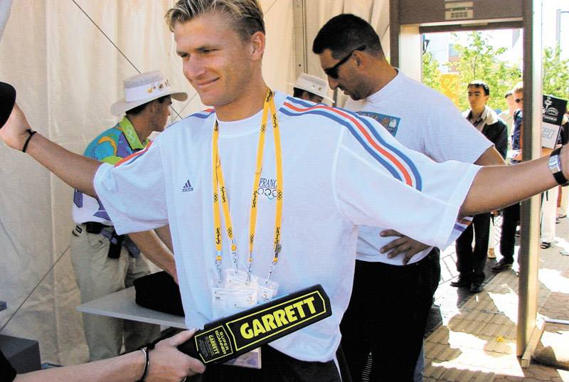 A person is being searched with a metal detector on their body. In the background, other people and a tent can be seen.