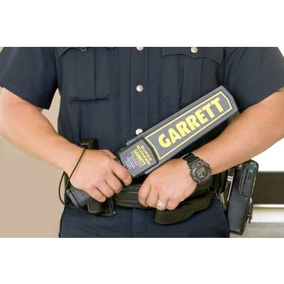 A security officer is holding a hand-held metal detector branded 'GARRETT'. He is wearing a black uniform with belt accessories.