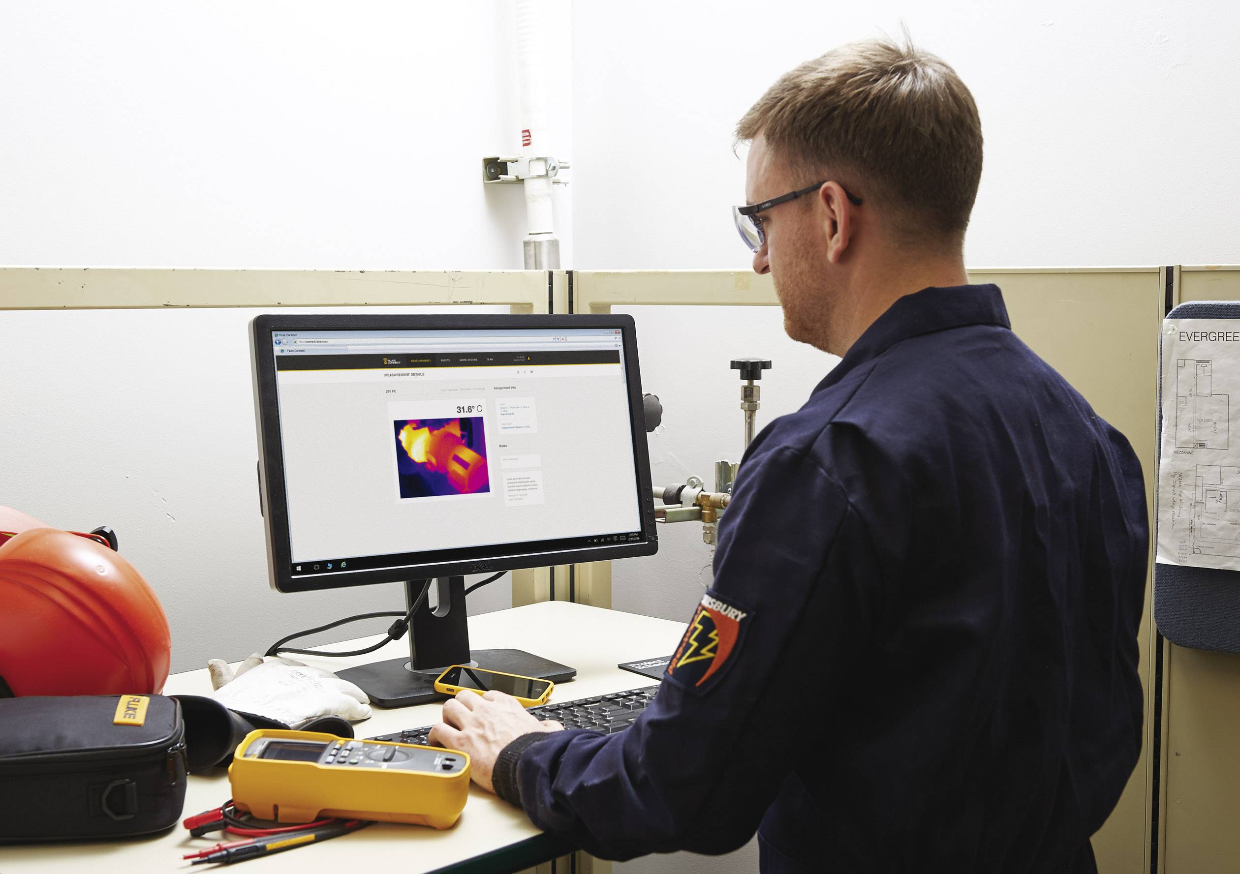A man is examining a thermal image on a computer screen in an office. Tools and a helmet are lying on the desk.