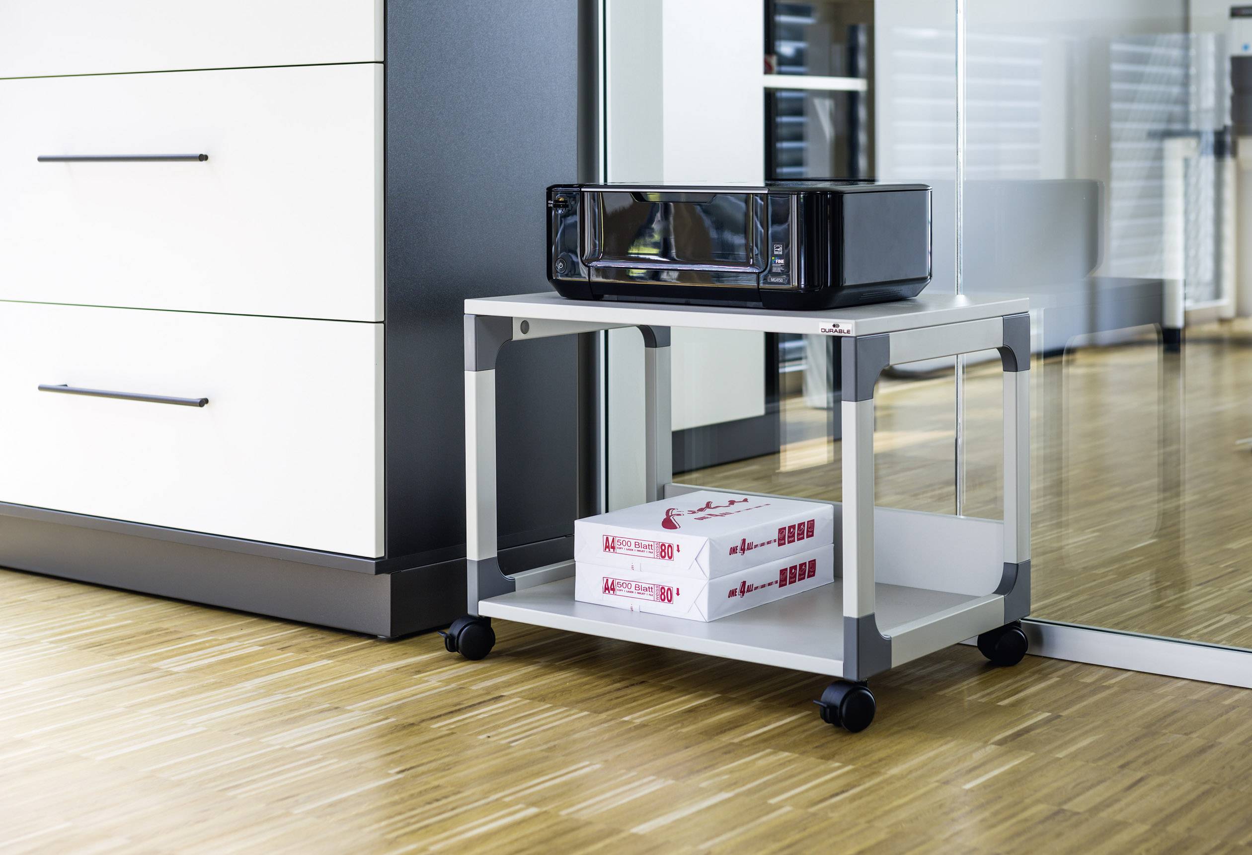 A printer sits on a mobile storage trolley in a modern office, with two stacks of printer paper underneath.
