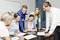 Six people in an office are discussing documents and diagrams. One person is pointing to a tablet while others listen attentively.