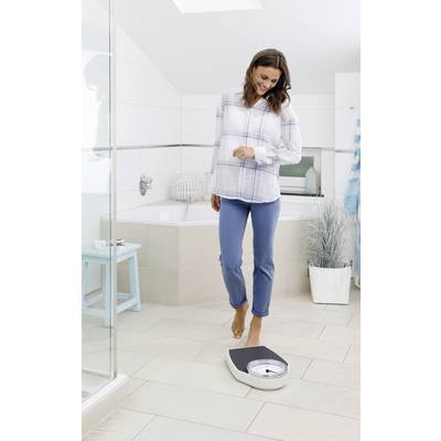 A woman stands barefoot on a bathroom scale, smiling down. The background shows a bright, modern bathroom setting.