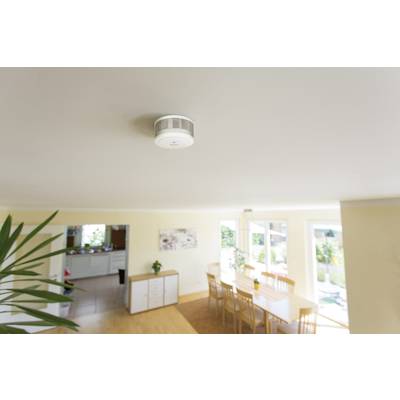 A smoke detector is mounted on the ceiling of a bright living room. Below it, a table with chairs, and large windows in the background.