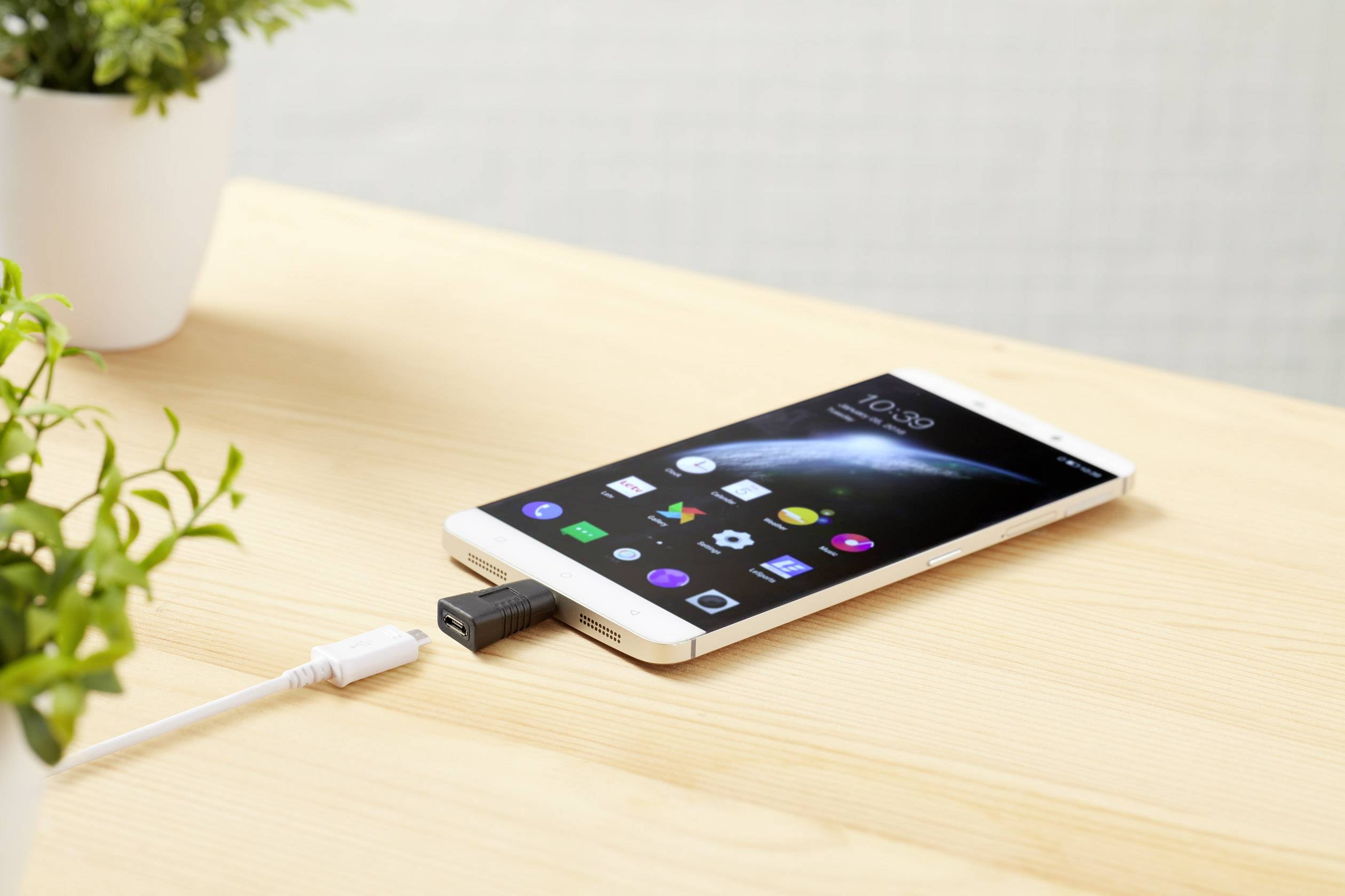A smartphone rests on a wooden table, connected by a USB cable. The display shows apps and the time. Plants are visible in the background.