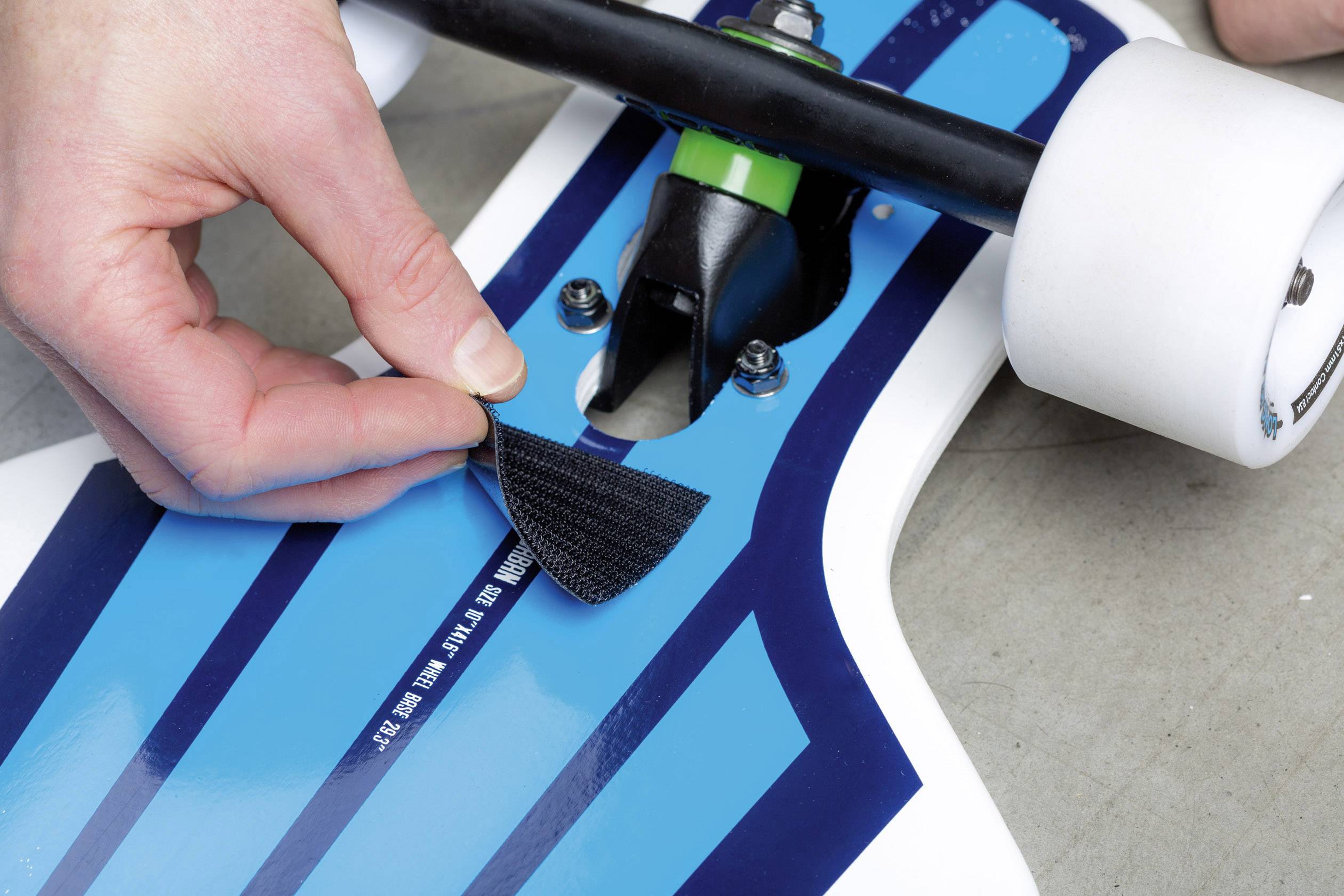 A hand is attaching Velcro to a blue and white skateboard deck near the rear truck.