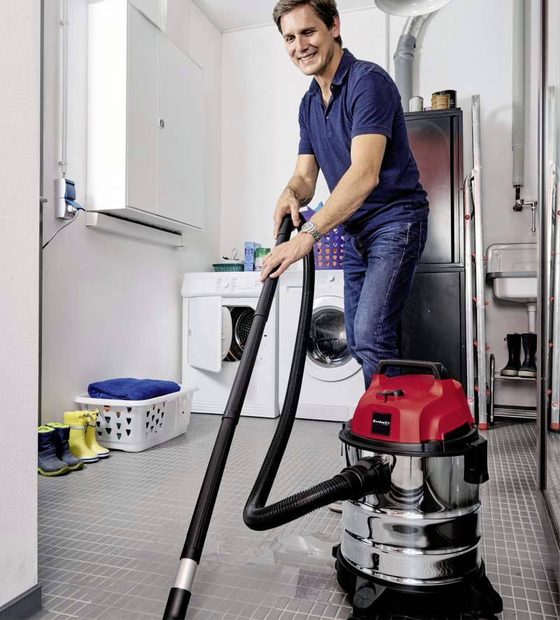 A man is using a wet and dry vacuum cleaner to clean the floor in a utility room. In the background, a washing machine and a laundry basket can be seen.
