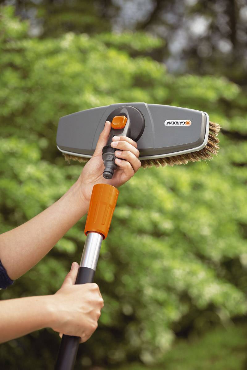 Two hands are holding a scrubbing brush head with an orange handle, outdoors against a blurred green background.