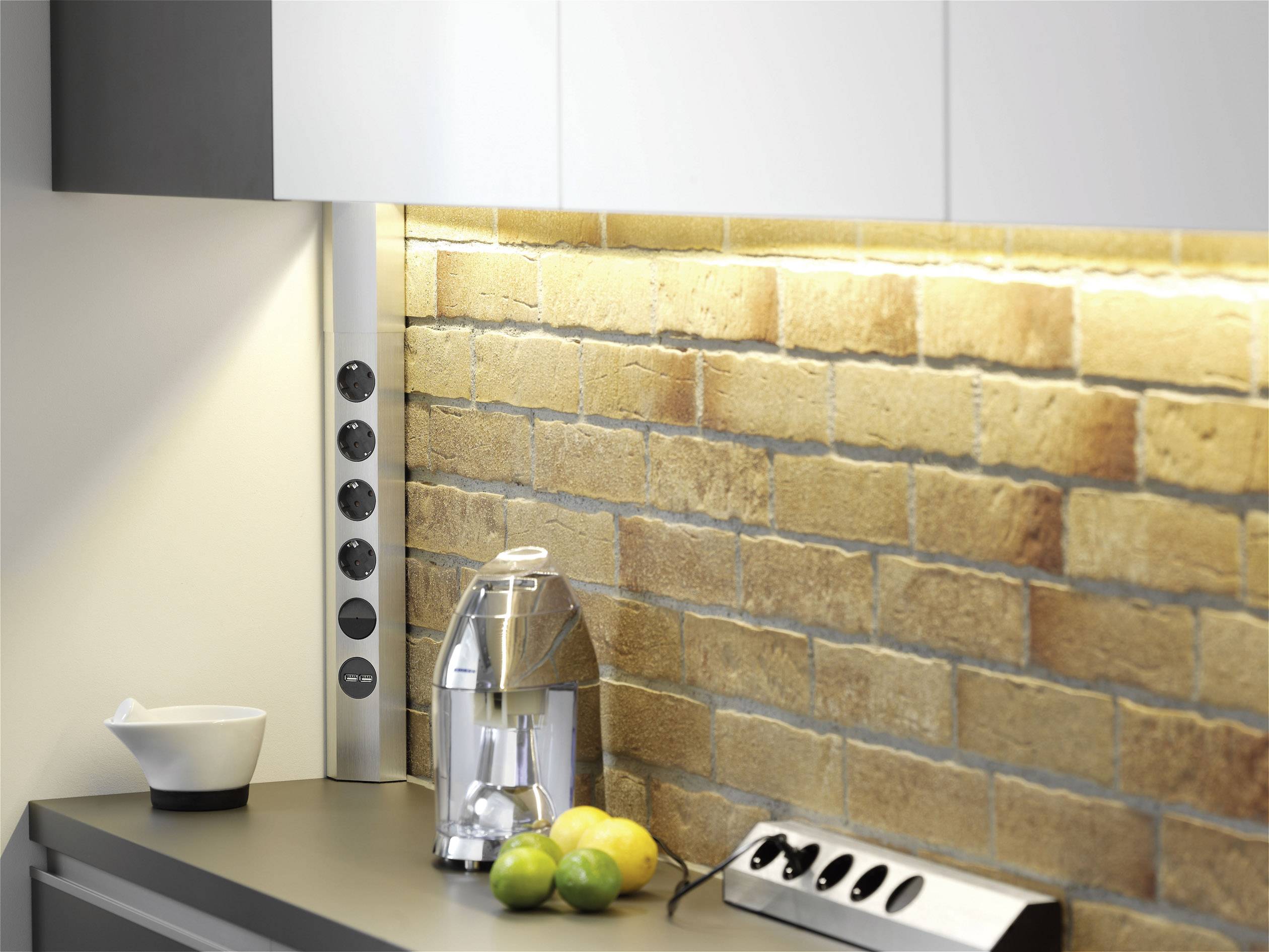 A modern kitchen worktop with a citrus press, lemons, and limes. The background shows an illuminated brick wall.