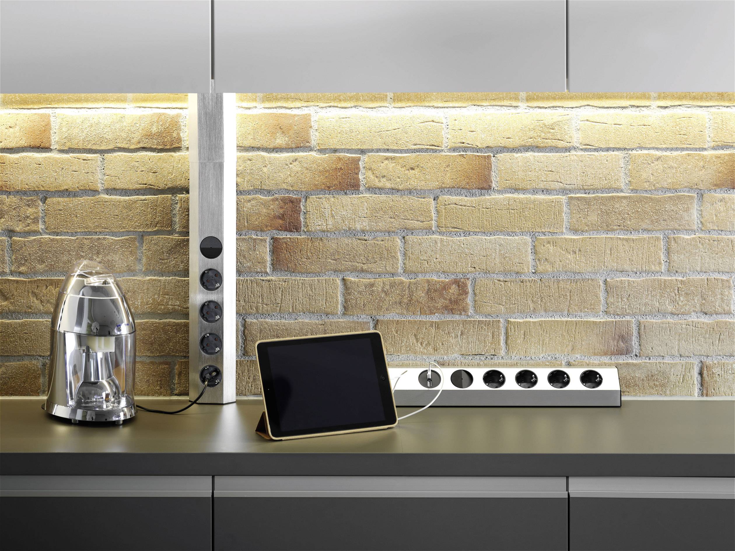 'Kitchen worktop with tablet and food processor. Wall rail with plug sockets, brick wall in the background.'