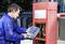 A man in blue workwear is using a portable diagnostic device to inspect a barrier control system outdoors.