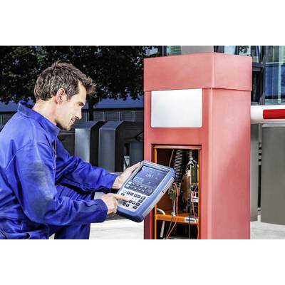 A man in blue workwear is working with a digital device on a red barrier control system. Buildings can be seen in the background.