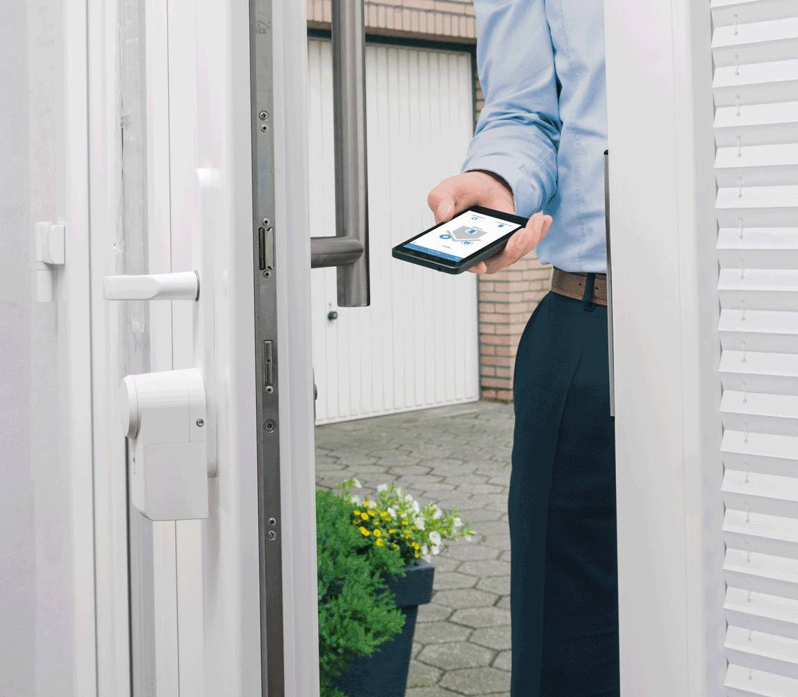 A person is holding a smartphone in front of an open door with a smart lock. Garages are visible in the background.