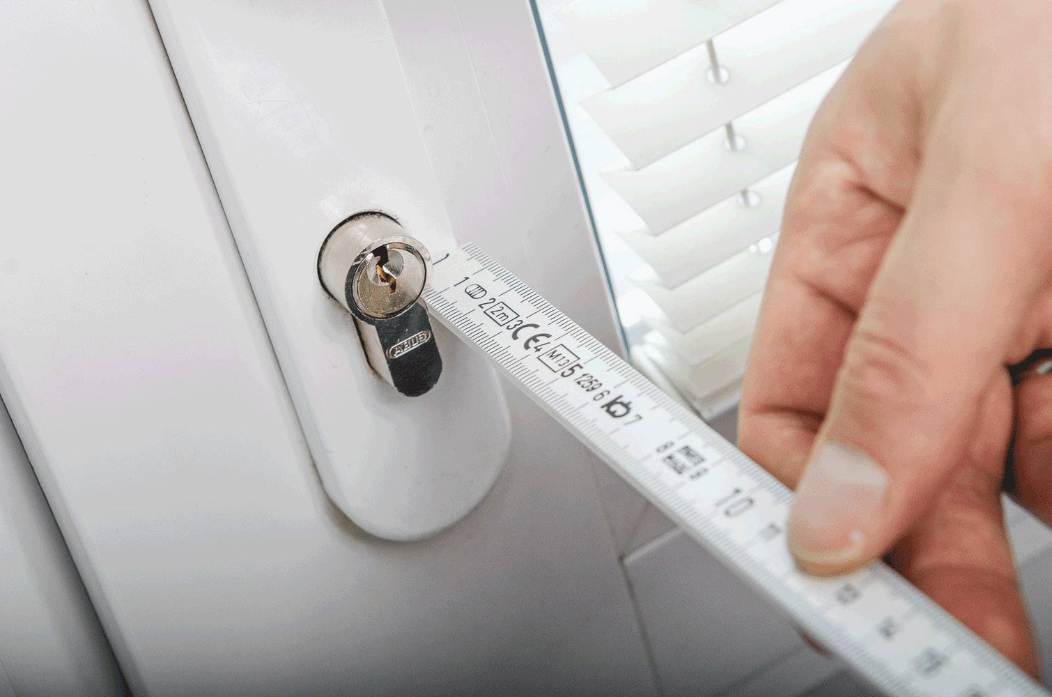 A hand holds a tape measure next to a closed window with blinds to measure the distance from the lock cylinder.