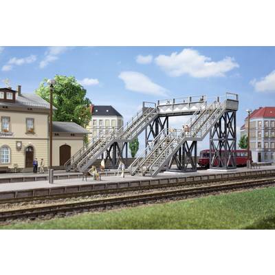 Model railway scene: A metal pedestrian bridge over railway tracks. People are standing and walking on the bridge. Buildings in the background.