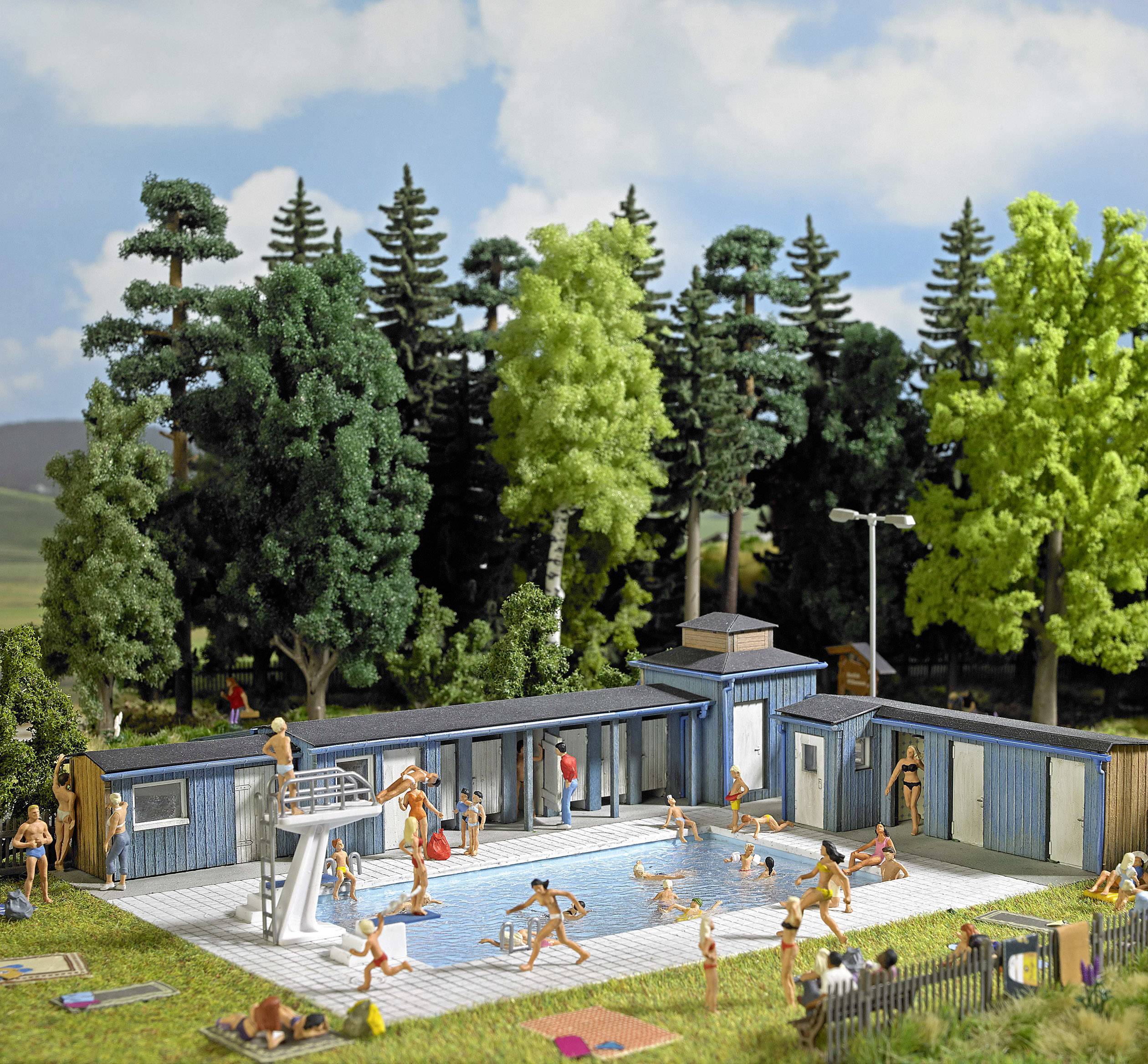 Miniature model of an outdoor swimming pool with people playing in the water or relaxing by the poolside. A wooded background frames the scene.