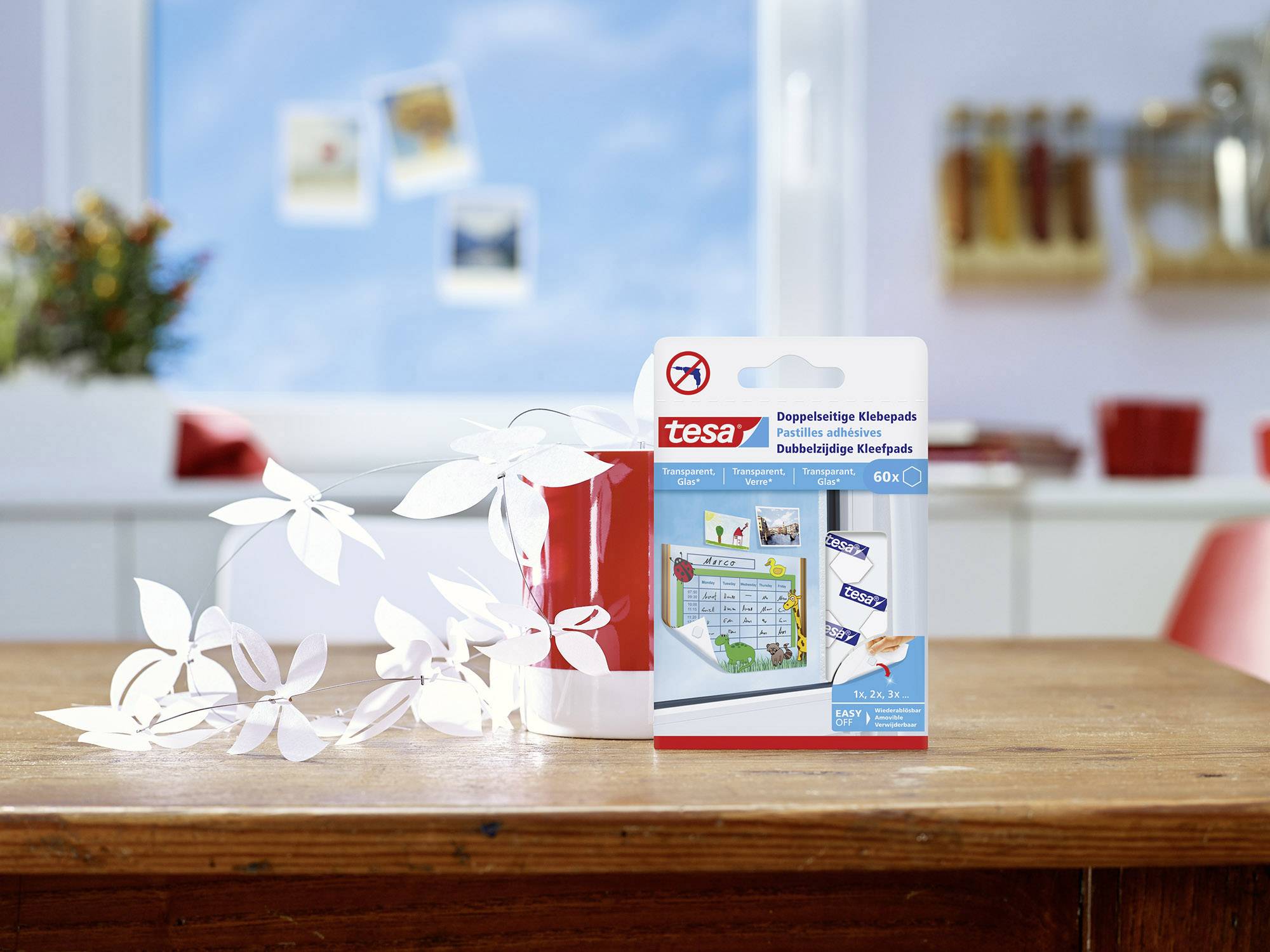 Packaging of double-sided tesa adhesive pads on a table, decorated with white paper sheets, with a kitchen in the background.