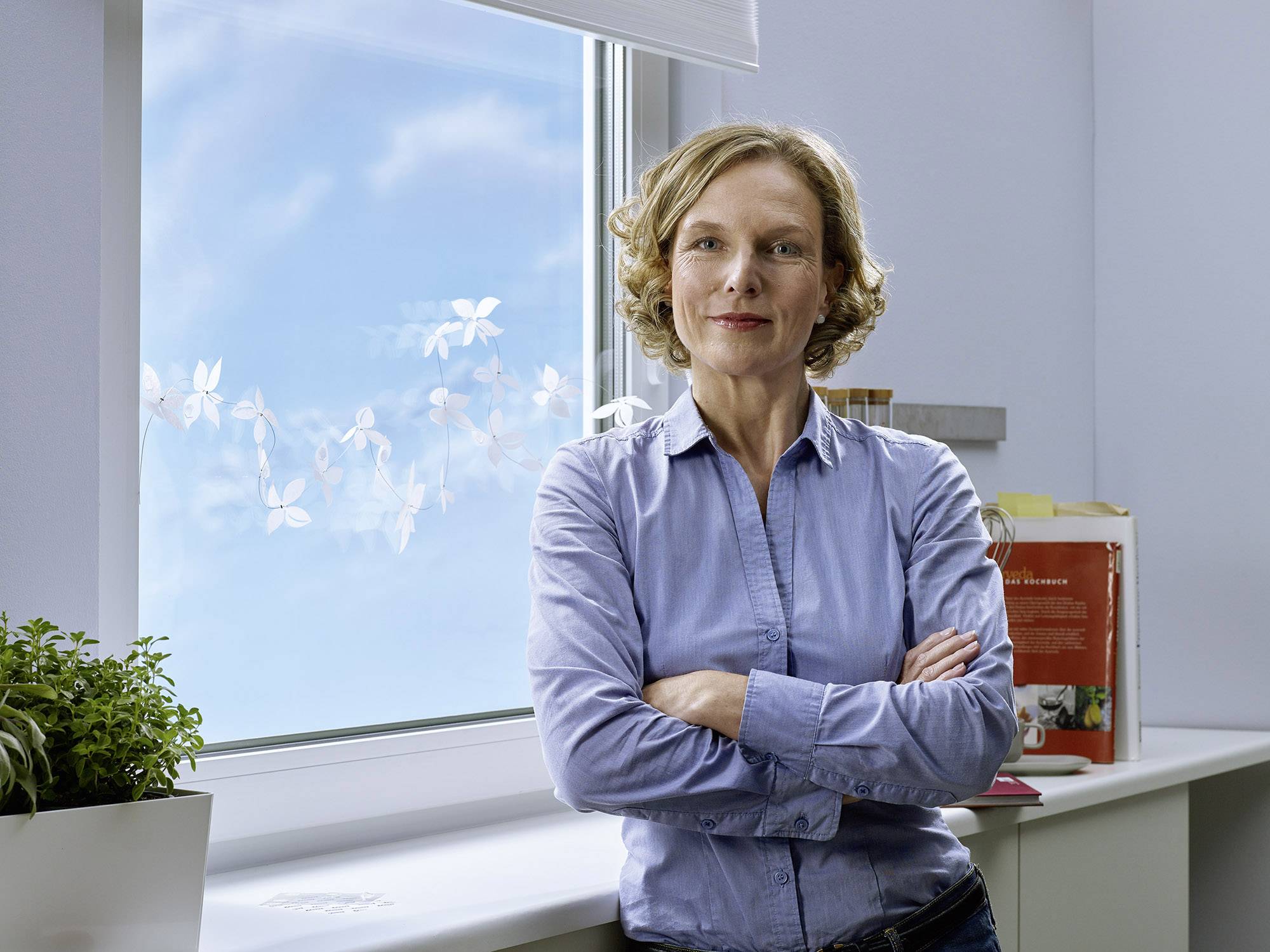 A woman in her middle years stands relaxed with her arms crossed in front of a window. A sunny sky is visible in the background.