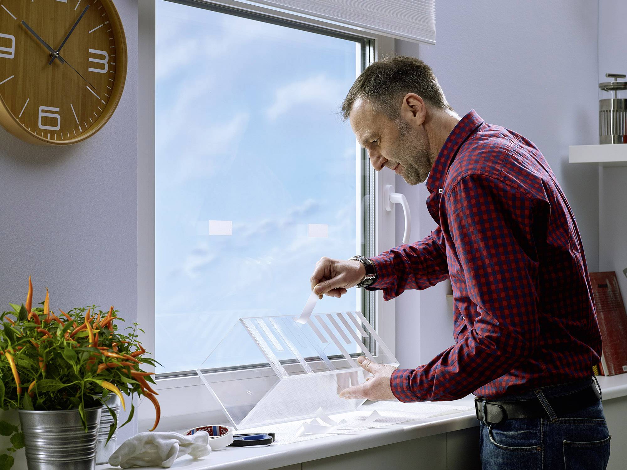 A man in a checked shirt is planting seeds in a small indoor greenhouse box on a windowsill. A bright room can be seen in the background.