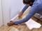 A man is fixing a white skirting board to a wall on a wooden floor. He is wearing a blue long-sleeved shirt and working carefully.