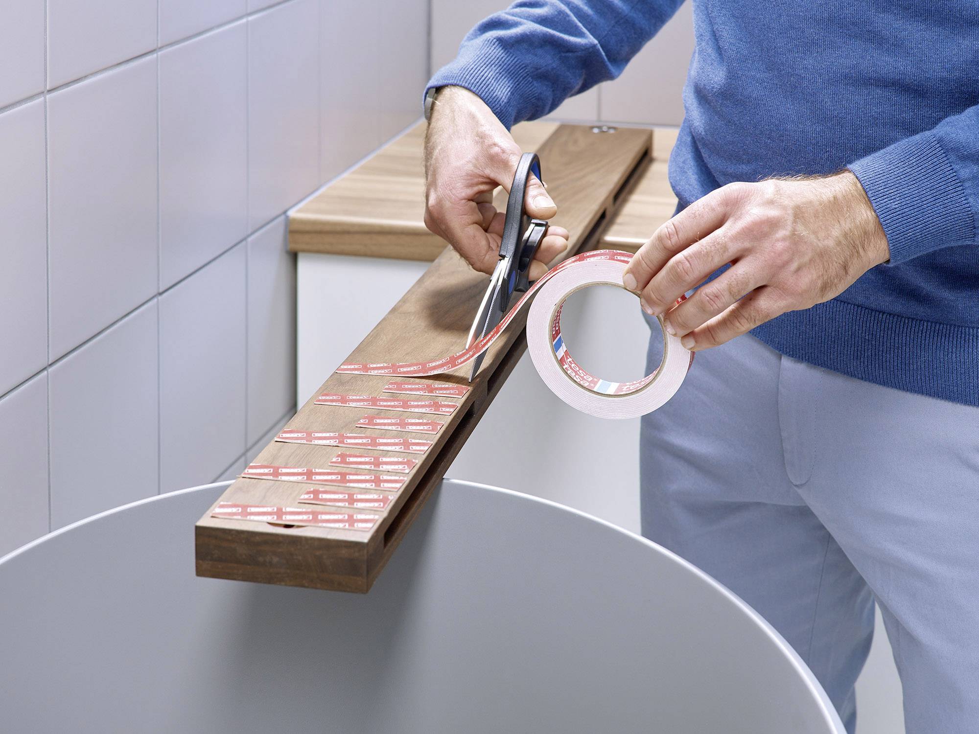 A person is cutting a piece of double-sided tape from a roll using scissors, ready to stick it onto a wooden board.