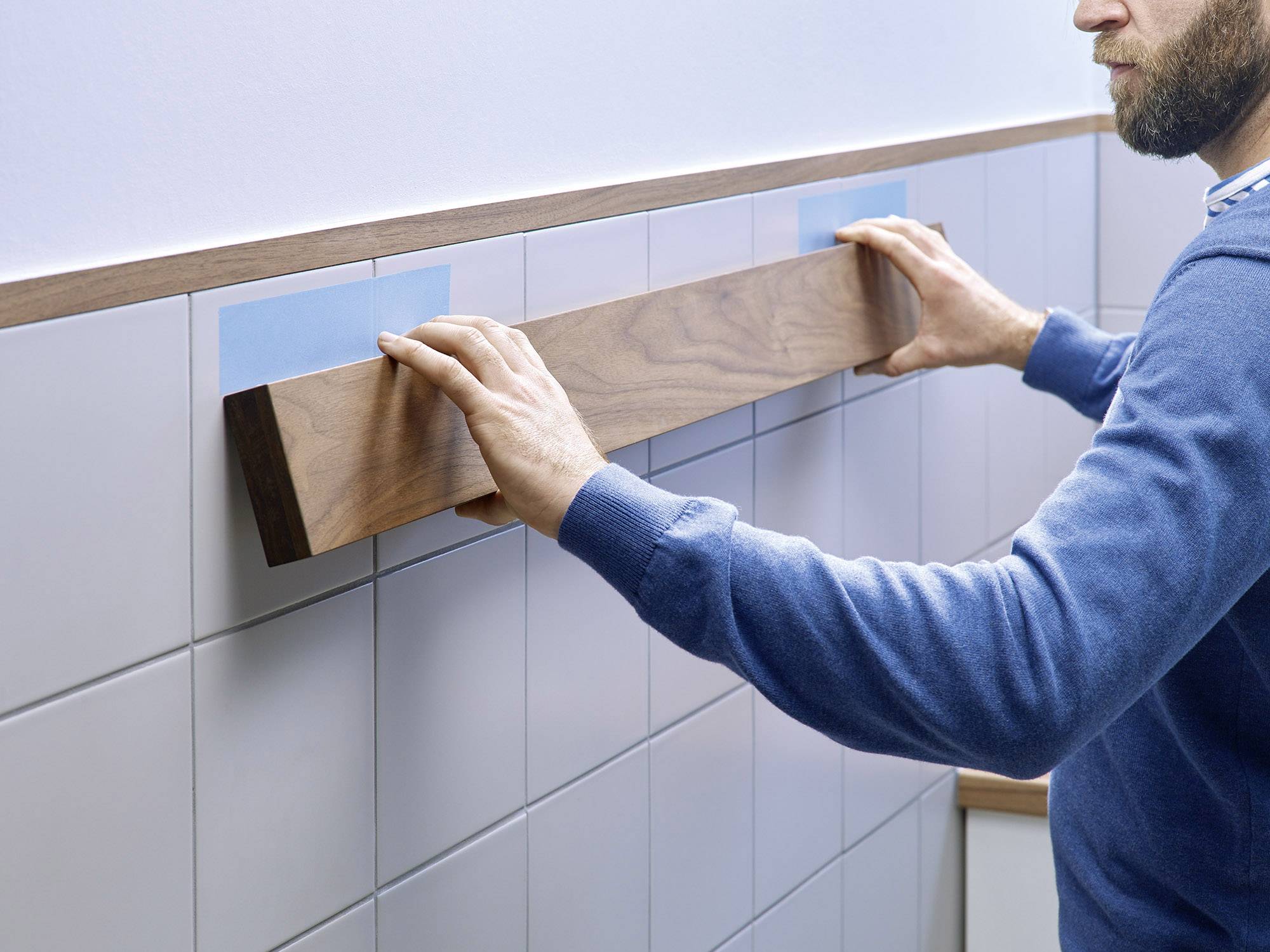 A person is attaching a long wooden board to a tiled wall with adhesive tape.